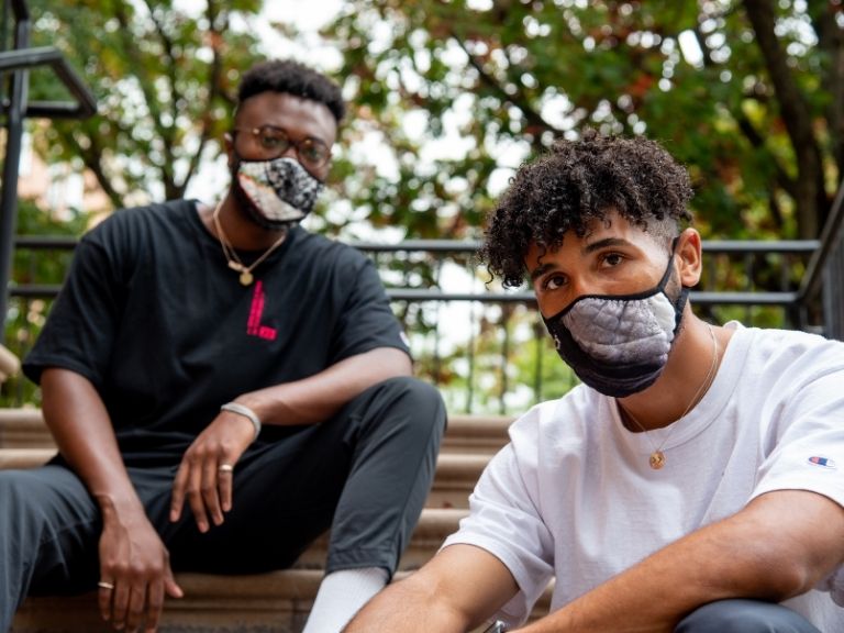 two male college students wearing face masks sitting on staircase together