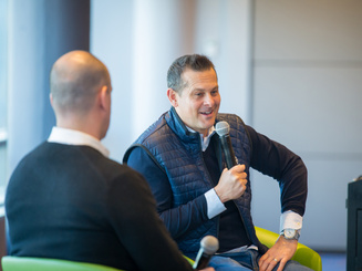 two people with microphones, sitting in chairs, giving a talk