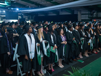 Undergraduate graduates stand together in regalia during commencement at Manhattan University awaiting the conferral of degrees.