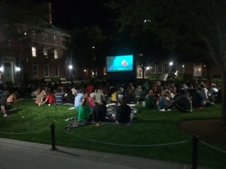 students sit outside watching a movie projected on a screen