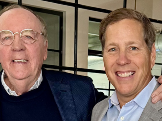 James Patterson and Patrick Leddin smiling together indoors, standing side by side in front of glass-paneled walls.