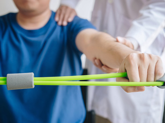Doctor physiotherapist assisting a male patient while giving exercising treatment on stretching his arm with exercise band in the clinic
