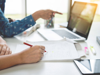 Student at table with laptop and person next to them with pencil and paper