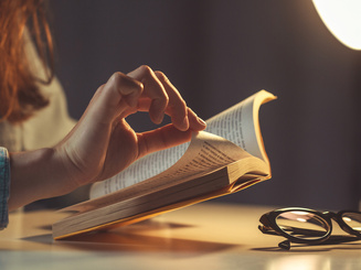 Person reading a book with a light and glasses on a table