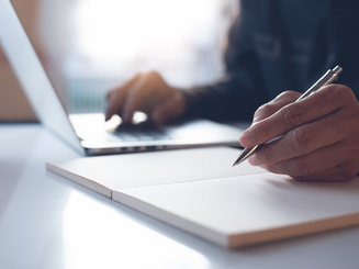 Person writing on a notepad while typing on a laptop