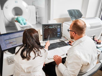 Back view of two doctors sitting, working at computer, examining MRI results. Specialists wearing white robes, consulting, woman pointing by pen, patient lying. 