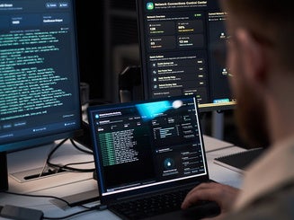 Vertical shot of person monitoring cybersecurity dashboards across laptop and desktop screens, analyzing network data and threat alerts.