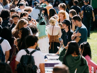 A diverse group of Manhattan University students and guests gathers outdoors around a table displaying clothing, with some individuals examining items and others conversing during an Open House.