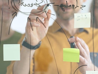 Business people colleagues brainstorming, writing strategy notes on glass board, collaborating with colorful sticky notes in creative workplace meeting in bright office