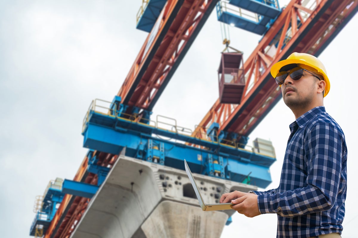 Engineer wearing a hard hat and sunglasses uses a laptop while inspecting a construction site with heavy machinery.