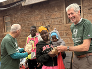 Brother Gus (left) and Brother Larry Schatz, FSC, distribute food to a Kenyan family during a service visit, standing outside a local community building.