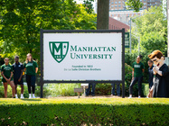 Students and the Jasper mascot stand near the Manhattan University campus sign displaying the new university name.