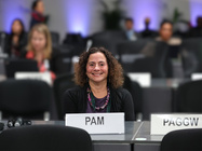 Pamela Chasek sits at a conference table with a name placard, smiling during a National Model UN session in a large meeting room.