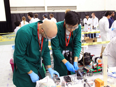 two students wearing lab coats and protective goggles work with chemistry equipment