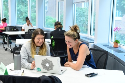 students seated at a table near windows, looking at a laptop screen together