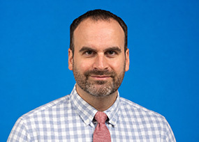 Portrait of Jonathan Gambier, Director of Transfer Recruitment, wearing a checkered shirt and tie against a blue background.
