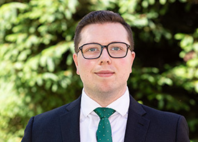 Portrait of Alex Plumacher, Associate Director of Undergraduate Admissions, wearing glasses and a suit with a green tie, standing outdoors.