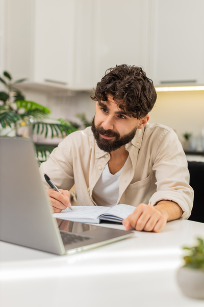 Person at home listens to online lecture on laptop writes notes in notebook
