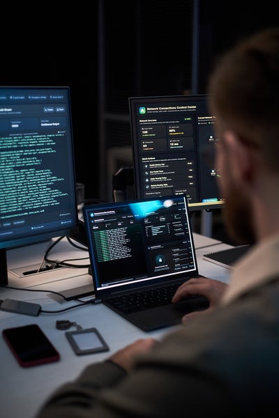 Vertical shot of person monitoring cybersecurity dashboards across laptop and desktop screens, analyzing network data and threat alerts.