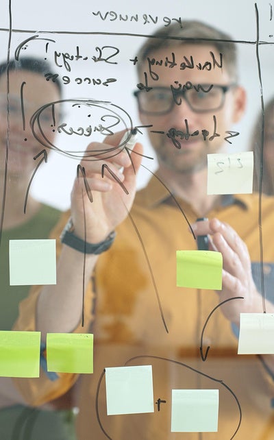 Business people colleagues brainstorming, writing strategy notes on glass board, collaborating with colorful sticky notes in creative workplace meeting in bright office