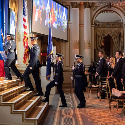 AFROTC color guard cadets march onto a stage carrying ceremonial flags during a formal event attended by guests and dignitaries.