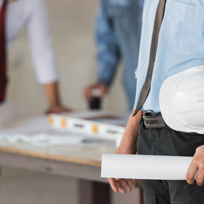 Young engineer holding a hard hat or white helmet and blueprint with worker at the construction site in background - teamwork concepts.