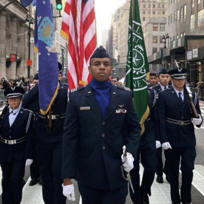 Manhattan University AFROTC cadets march in formation carrying American and Manhattan University flags during a New York City parade.