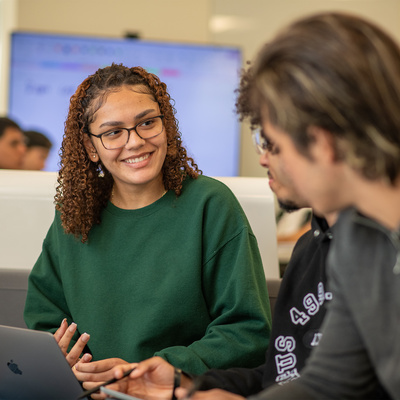 Manhattan University students in a classroom smiling and talking in front of a laptop